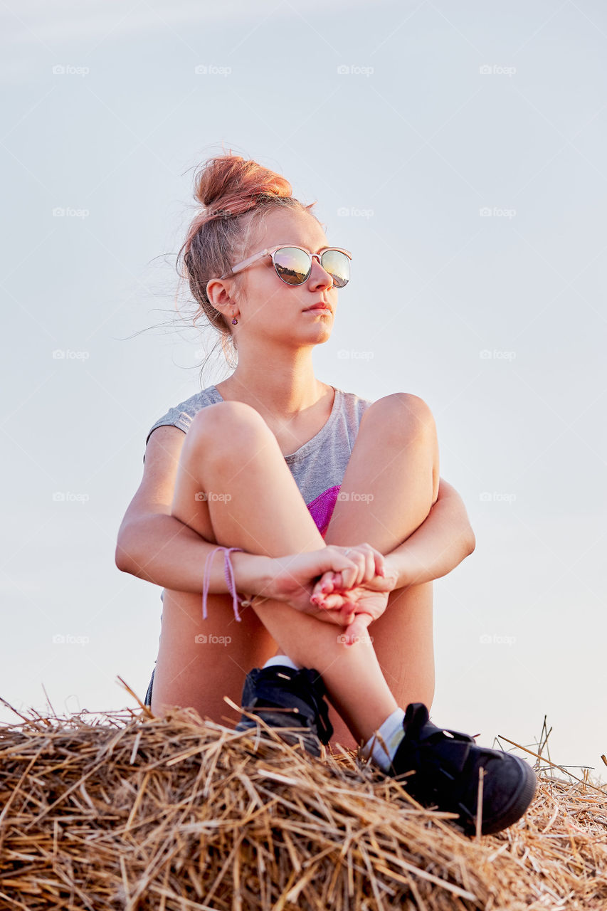 Teenage girl sitting on a hay bale at sunset relaxing while spending summertime in the village. Candid people, real moments, authentic situations