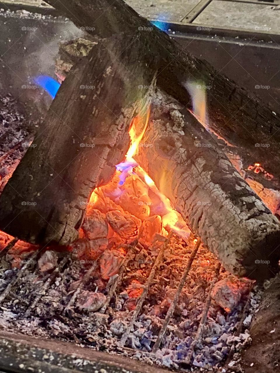 This is a photo of a campfire with burning logs. The embers and flames show a mix of red, orange, and blue colors, indicating the high temperature and presence of different chemicals.