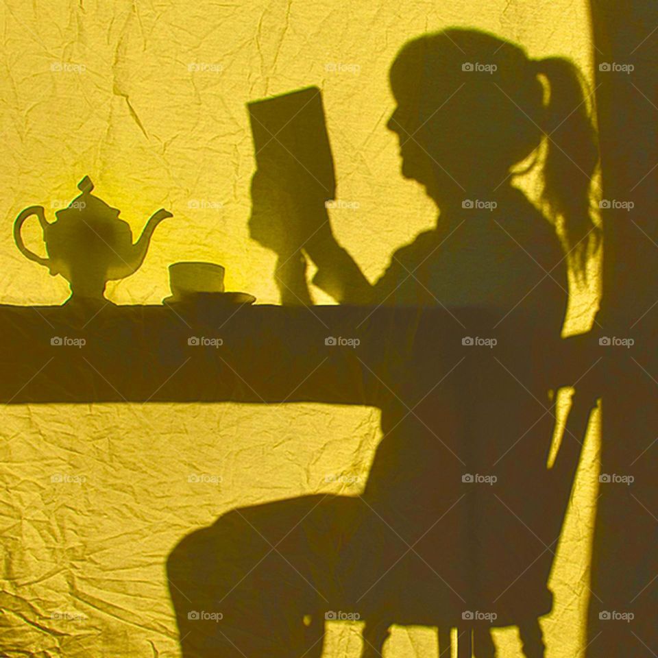 Silhouette of woman reading along with a pot of tea and a cup on a saucer