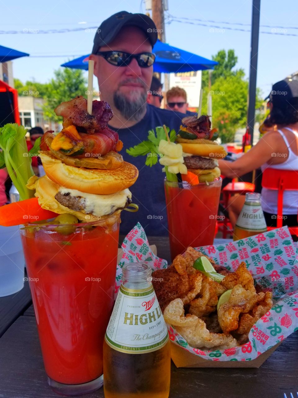 Happy man enjoying brunch time with amazingly loaded bloody mary cocktail complete with garnish stack with ice cold shorty Miller high life bottle sidekick and crunchy spiced homemade pork rinds on patio on sunny summer day.