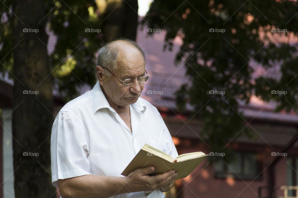 An elderly man of retirement age with wrinkles sits on the grass in the park and reads a book.