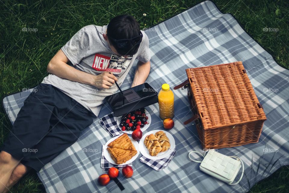 One young caucasian guy in shorts and a T-shirt lies reclining on a bedspread with a wicker basket, croissants and fruits in plates, browsing social networks in a tablet, relaxing in a clearing in a city park on a summer day, close-up from above.
