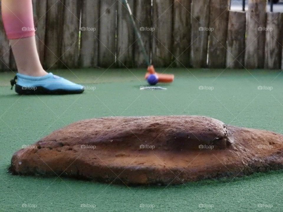 Mini golf playing low angle showing shot in the hole with blue water shoe with brown obstacle and a wooden fence in the background.