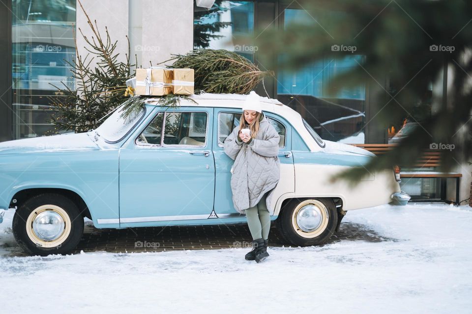 Young woman blonde girl in warm clothes with cup of coffee in hands near retro car decorated Christmas tree in winter street