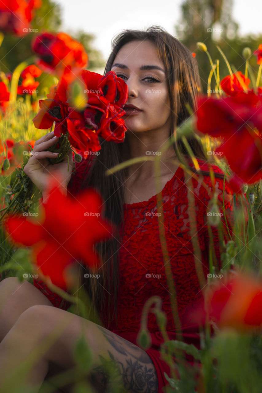 Beautiful woman with bouquet of poppies sitting in red dress enjoy a poppy field
