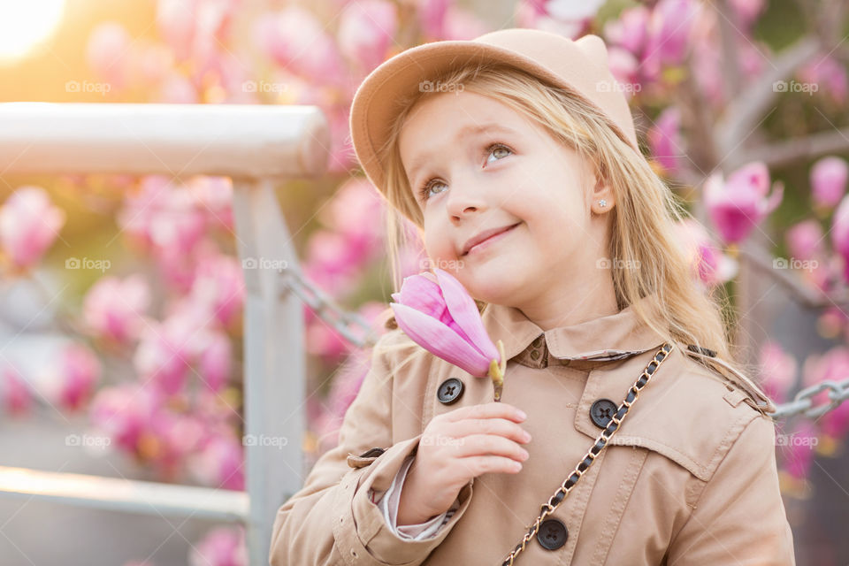 Portrait of cute stylish little girl with blonde hair which holding pink blooming magnolia flower 