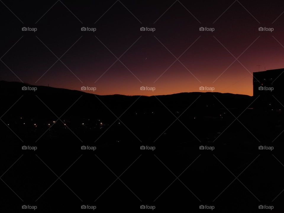 A beautiful evening landscape right as the sun is setting down, with pretty pink and orange colours and no clouds over the hills, with the moon showing in the sky