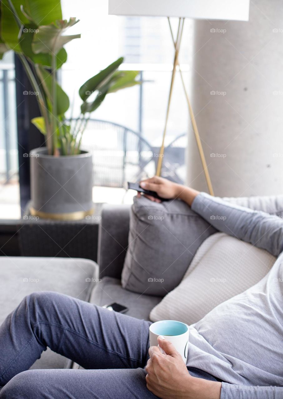 "The person is sitting comfortably on a gray sofa with a cup in his lap.
Large houseplants look out of focus."