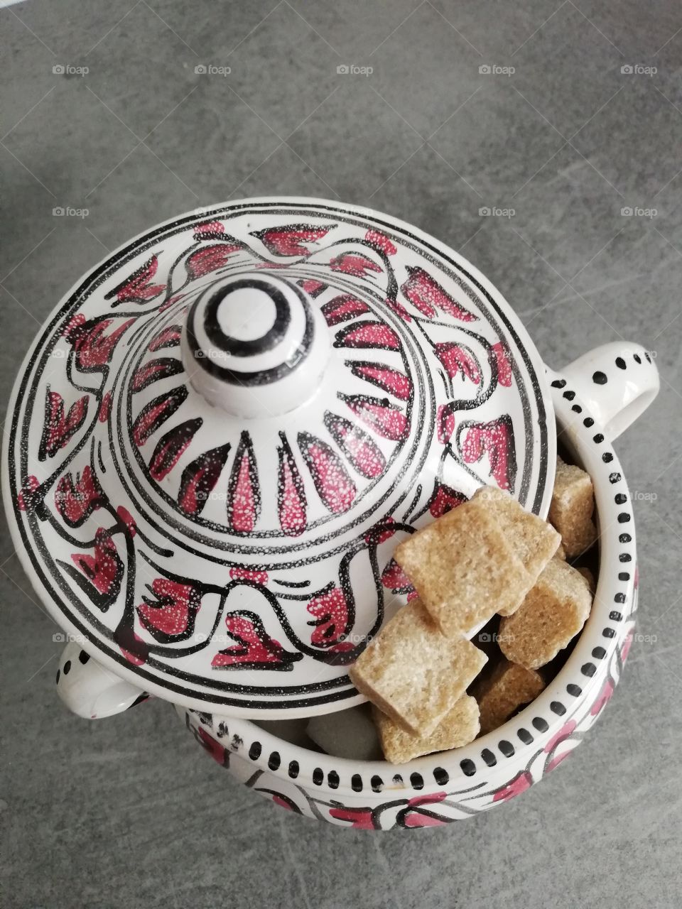 Multicoloured and patterned sugar container with handles and a striped top is full of white and brown pieces of sugar