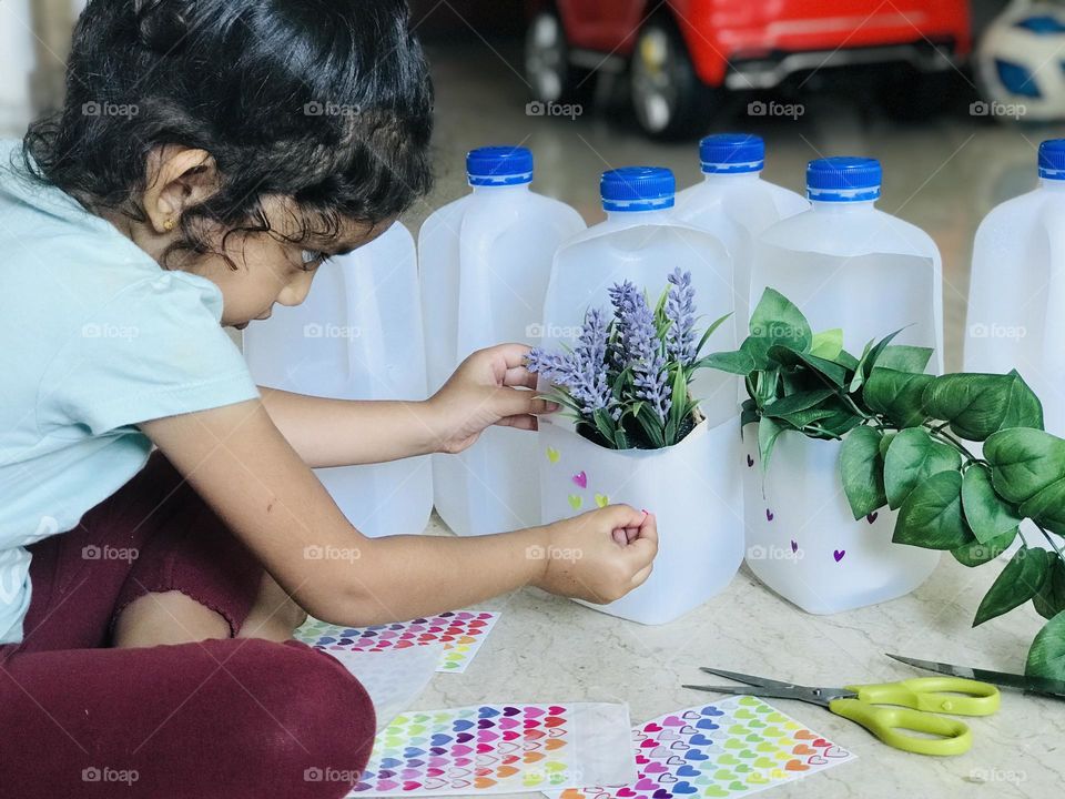 A girl doing craft with leftover plastic milk bottles. It converted into flower pots and pasting stickers on the pots.