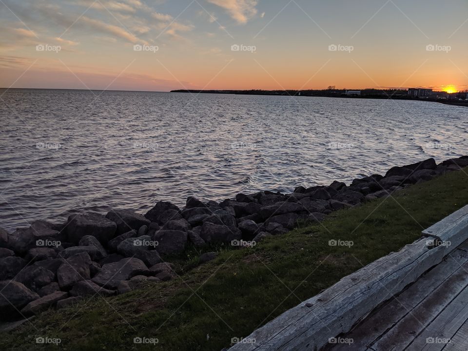 waterfront at sunset, wooden path, rocks, and grass in foreground.