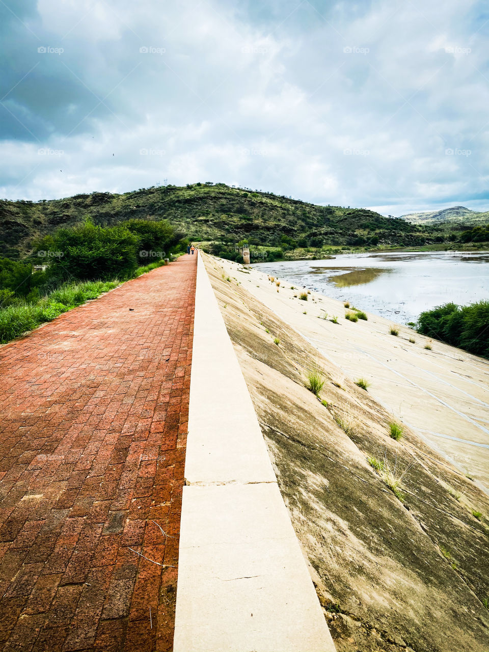 Concrete wall on the walls of the dam with muddy rain water, green vegetation and mountains on a cloudy day.