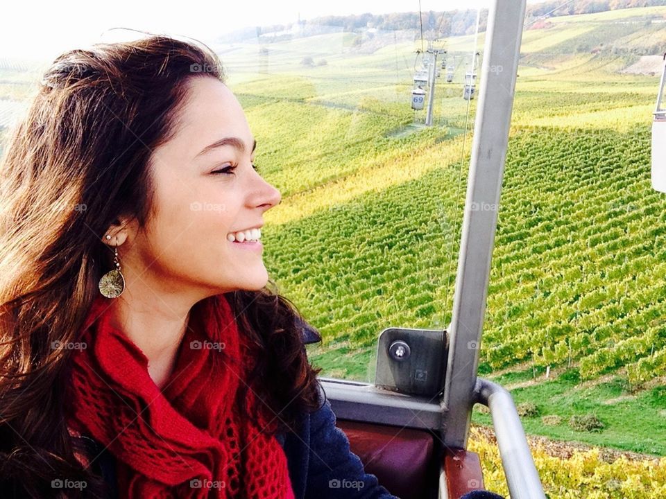 The face of a smiling woman is in the focus of the photo together with her red encharpen. She is sitting on a red bench of a cable car overlooking the grape plantations, vineyards