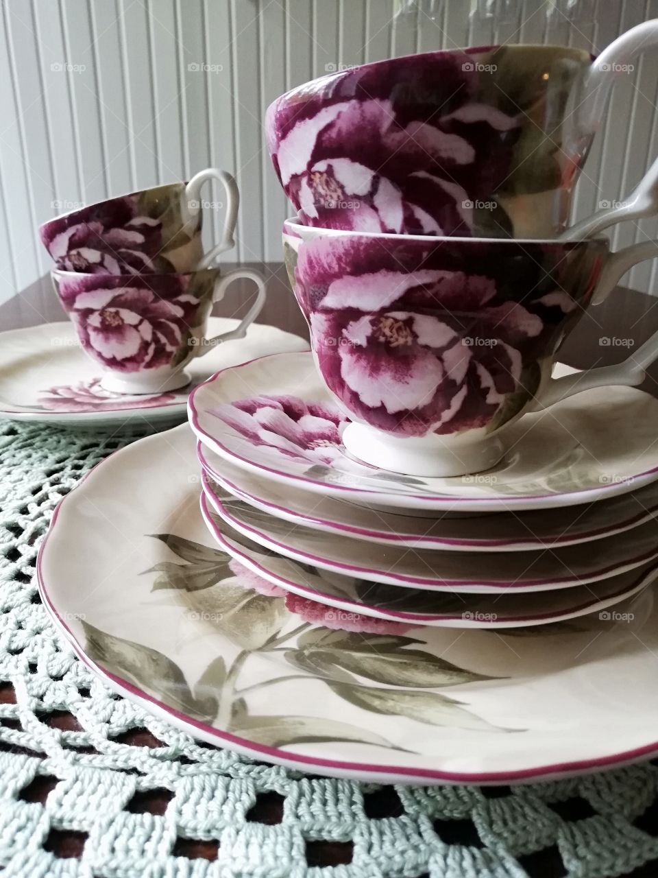 Patterned coffee cups on the green lacy table cloth on a brown table. Flowers are on the surfaces and a stripe on the edges of the plates of two different sizes, behind glasses on the wall.