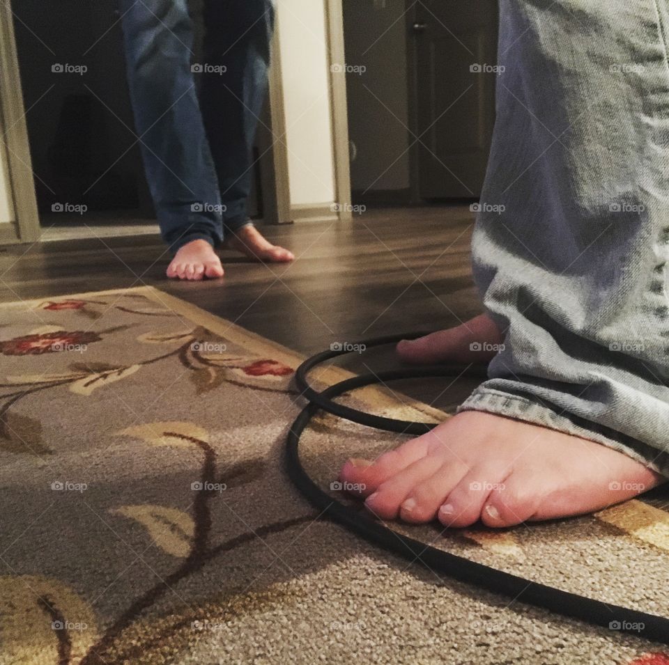 The feet of two people who are wearing blue jeans, one light, one dark, both standing on a dark wood floor with a blue, white, and green floral rug and a musician’s auxiliary cable.