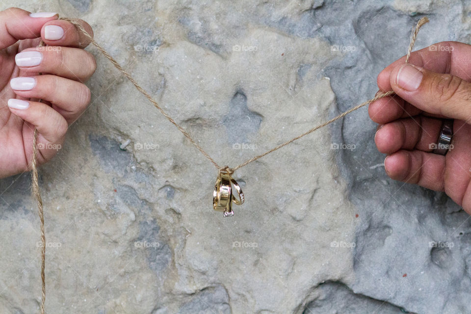 Two hands holding a string tying gold wedding bands together in front of a stone wall