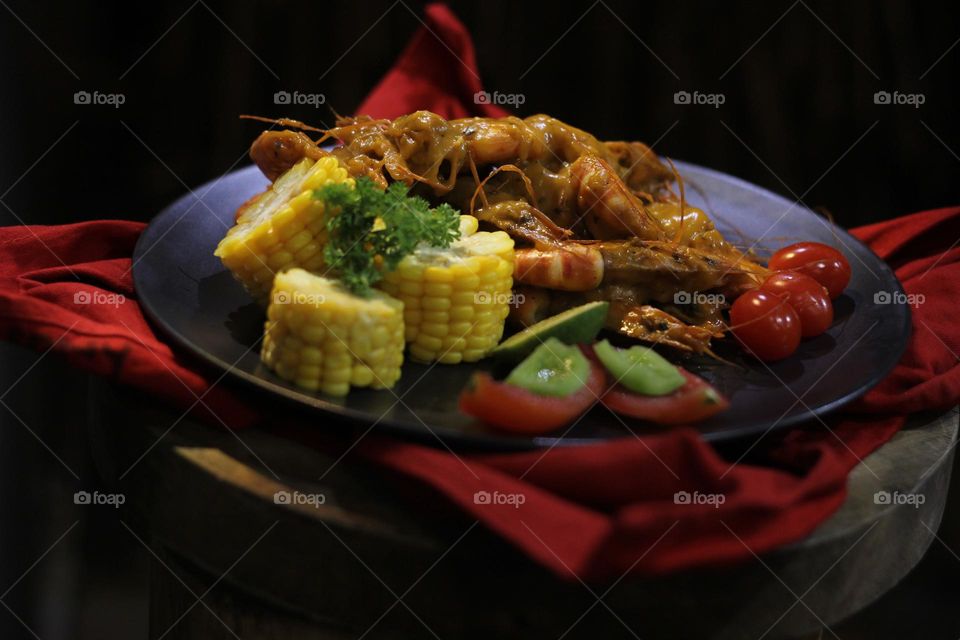 Asian food balado fried prawns with corn chunks, lemon slices and tomatoes served on black plate with red cloth bottom and wooden coaster. Asian style street seafood.