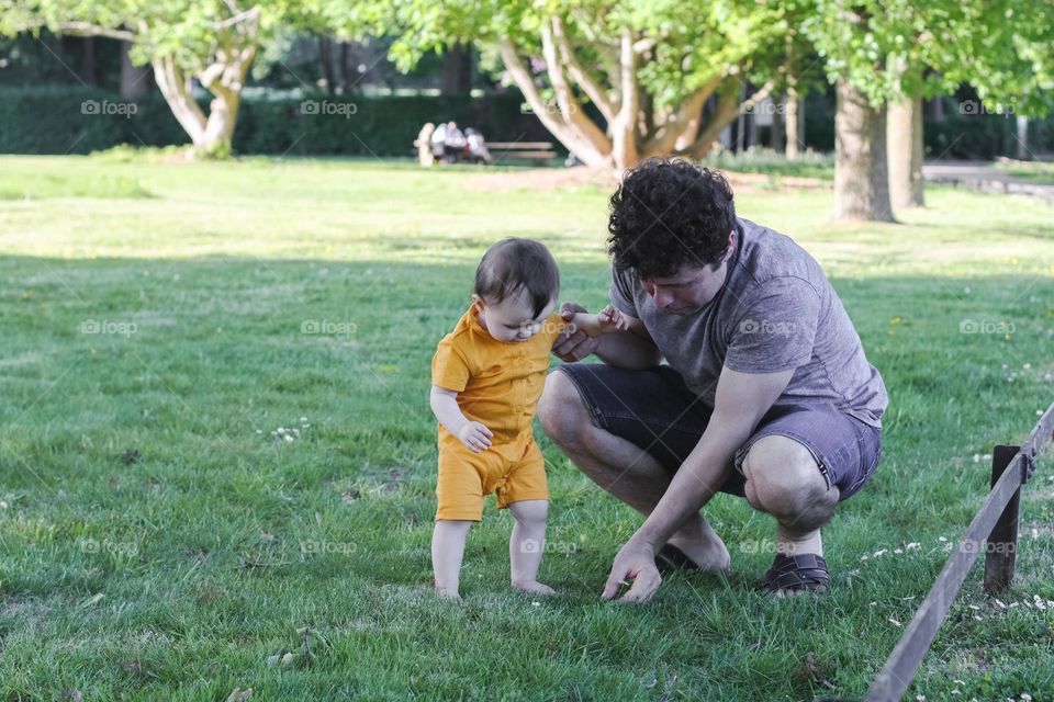 Portrait of a young caucasian man tearing a chamomile for his little baby sitting on a lawn in a public park behind a small wooden fence with copy space on the left, close-up side view.Dads concept, outdoor walk, family vacation.