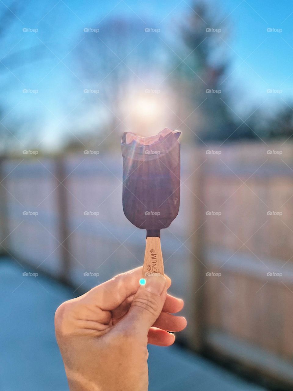Eating an ice cream bar in the sun, woman’s hand holding a Magnum Ice Cream bar, eating a Magnum ice cream bar, smartphone food photography, delicious desserts eaten outside, sunshine coming through bite on ice cream bar, Magnum ice cream brand