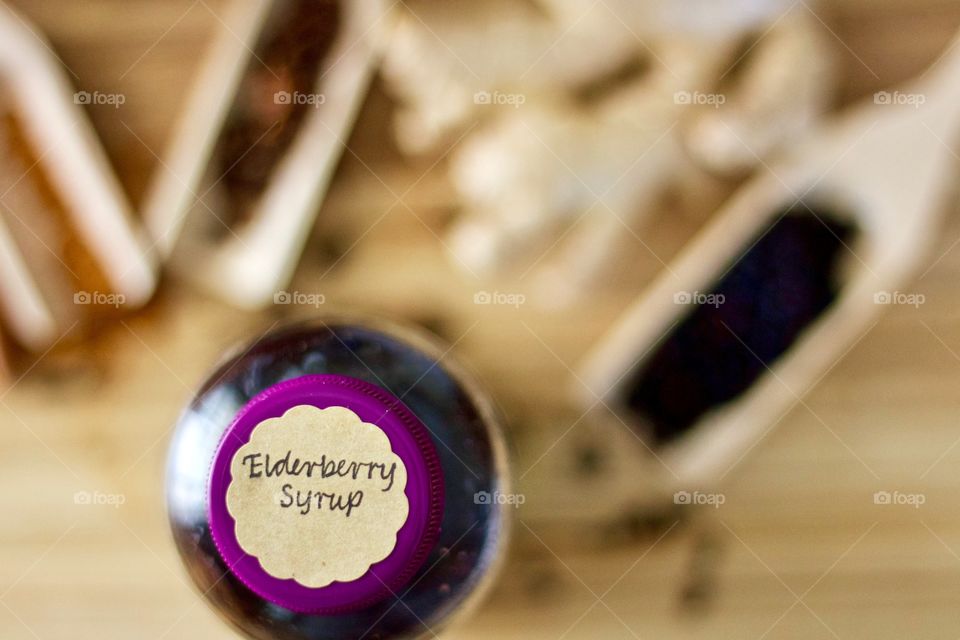 Overhead view of homemade elderberry syrup in a glass bottle with a labeled purple lid, blurred background including whole and ground spices in wooden scoops on a wooden surface 
