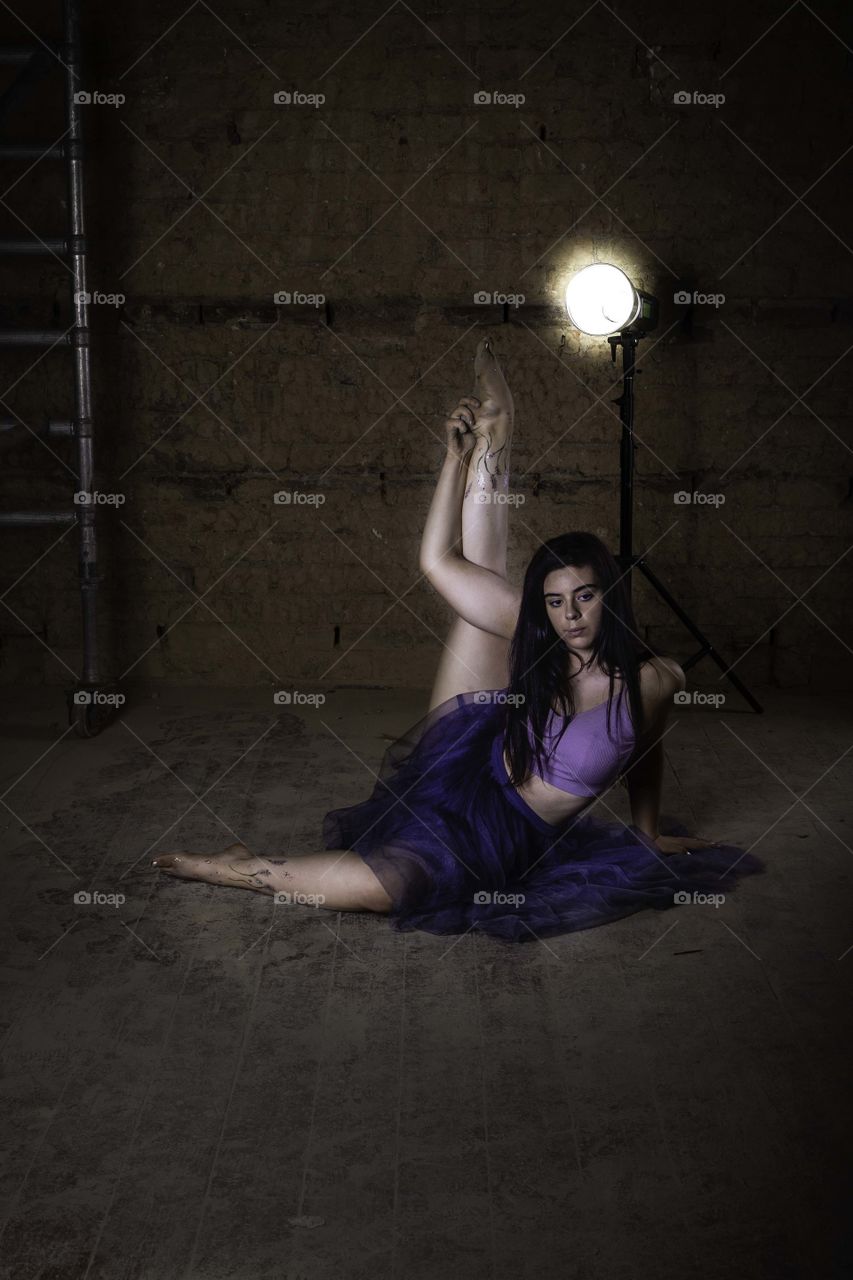 Female dancer stretching. She is holding her outstretched leg. Lit by light behind her wearing a crop top. Tattoo on her leg