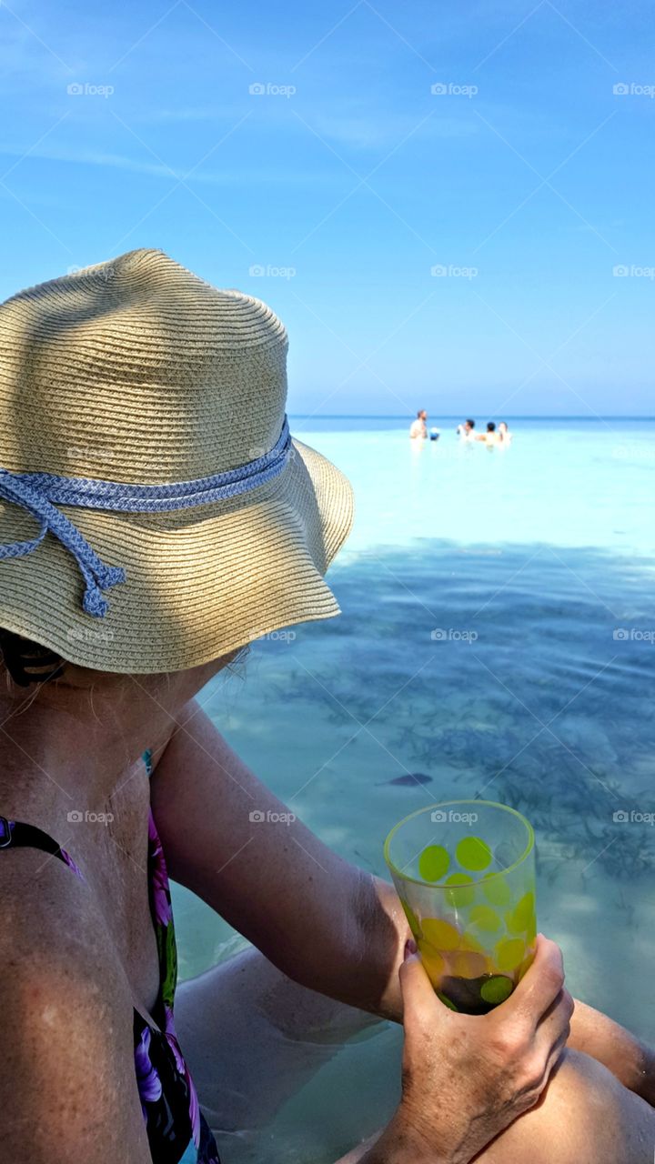 A woman on a beach in a hat watches people play in the ocean.