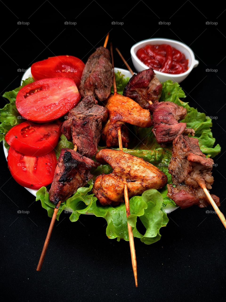 A white plate on which lie: green lettuce leaves, pieces of fried meat on a skewer, roasted wings on a skewer, chopped red tomatoes.  In the background is a white gravy boat with red ketchup.  Black background