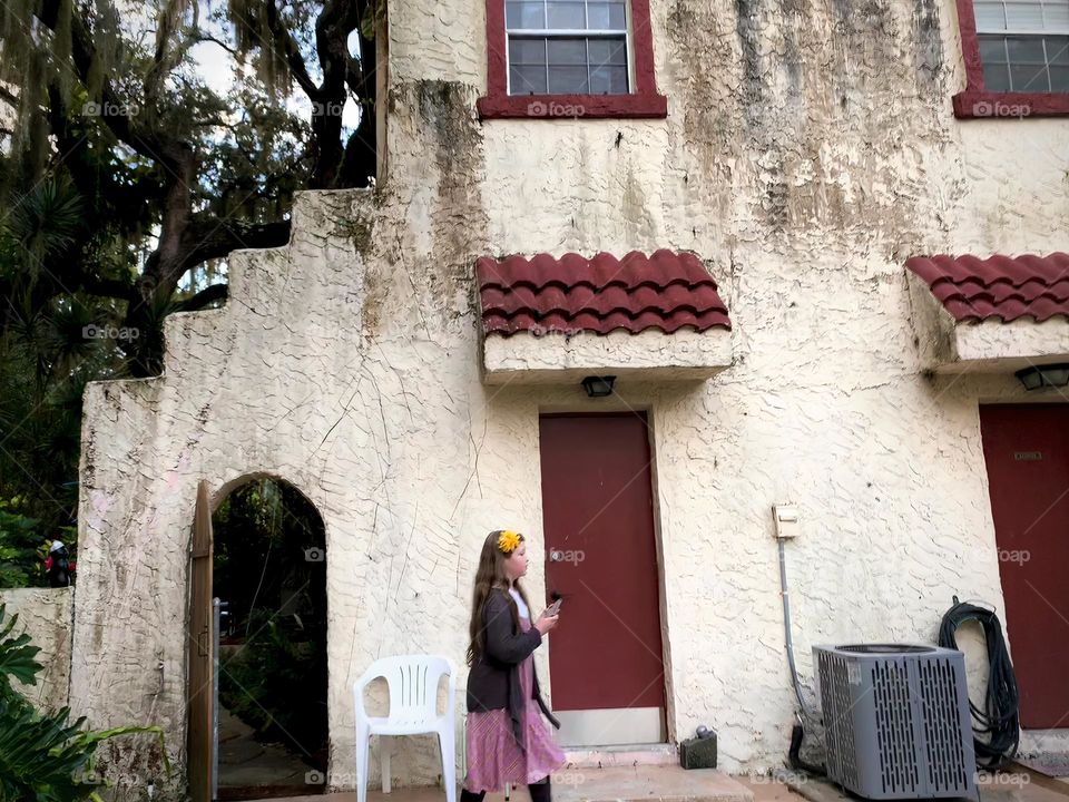 White and red spanish old style architecture residential large house built in the early 1900s. Doorway with antique style wood door in a stairway wall with white chair by a red door and window seen from tropical plants, live oak trees with child walk