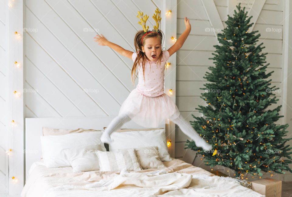 Cute little girl in pink dress jumping on bed in room with Christmas tree. Child in rim with deer horns in cottage house in the christmas time, happy New Year