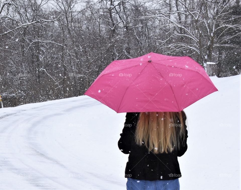 Pink Totes umbrella held by a lady with long blonde hair walking down a country road during a snow storm 