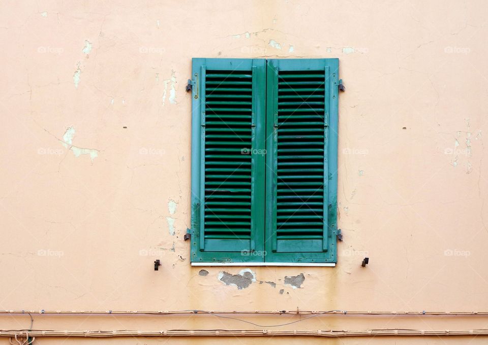 Full frame shot of pink colored weathered building exterior with window behind closed green shutters and fixed cables in Empoli, Italy.