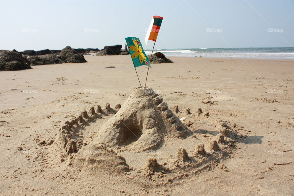 A sand castle built on sea shore. The castle is having victory flags on it.