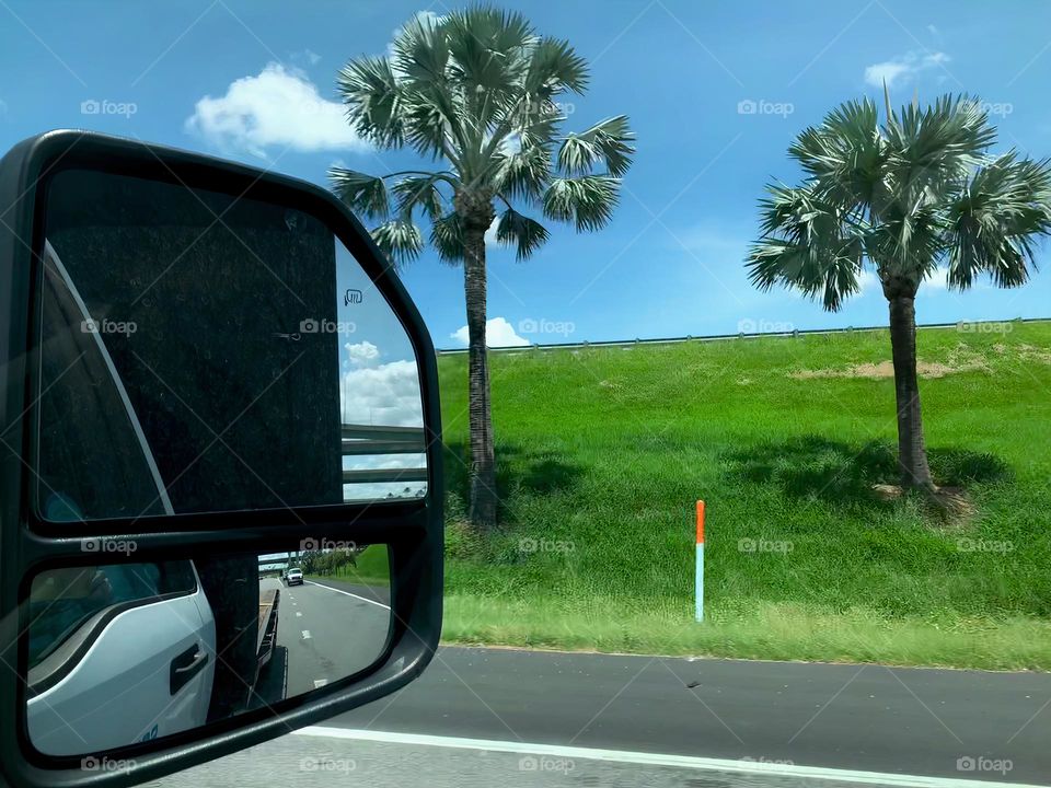 Commuting: transporting people to get heavy material from one place to another seen from visor passenger side mirror from a flatbed truck during a nice day with clear blue sky and a little bit of clouds with green grass and palm trees on the highway.