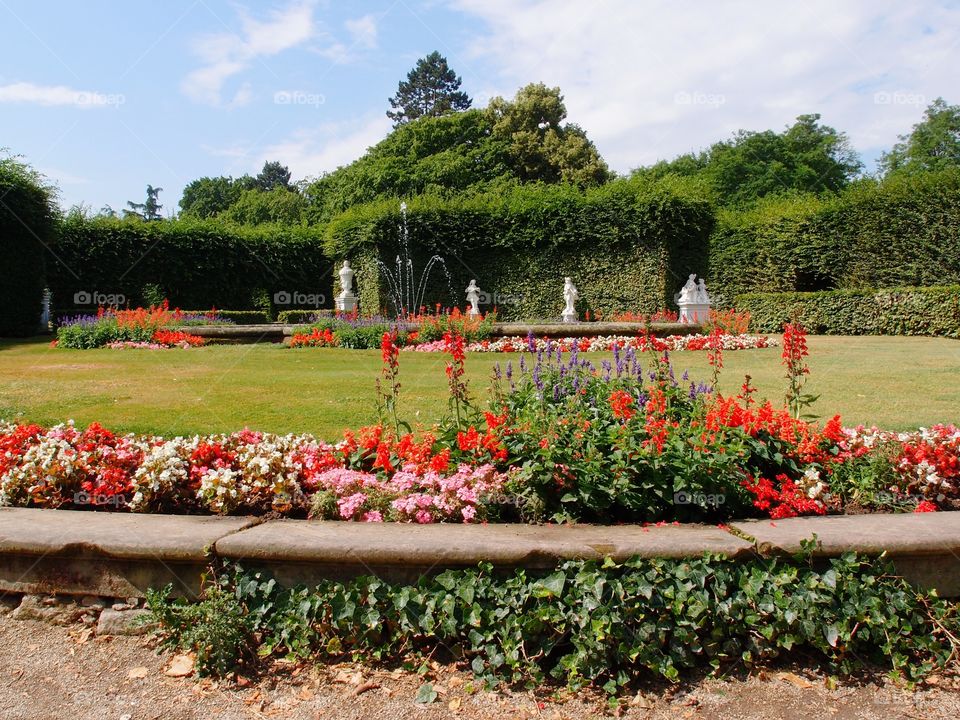 Bright red, pink, purple, and white flowers with a beautiful fountain in the background along with statues and thick hedges in a public European park on a sunny summer day. 