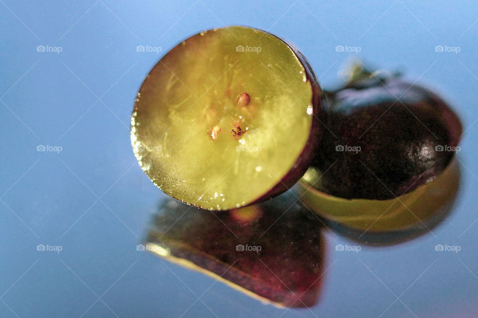 A macro of a blueberry cut in half with 1/2 the berry, the greenish/pinkish flesh with seeds facing up. The other 1/2 the berry flesh is facing down, with the bluish/purplish berry skin exposed. Both reflect opposites sides of the berry in the mirror