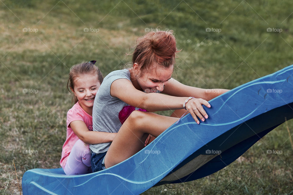 Teenage girl playing with her younger sister in a home playground in a backyard. Happy smiling sisters having fun on a slide together on summer day. Real people, authentic situations