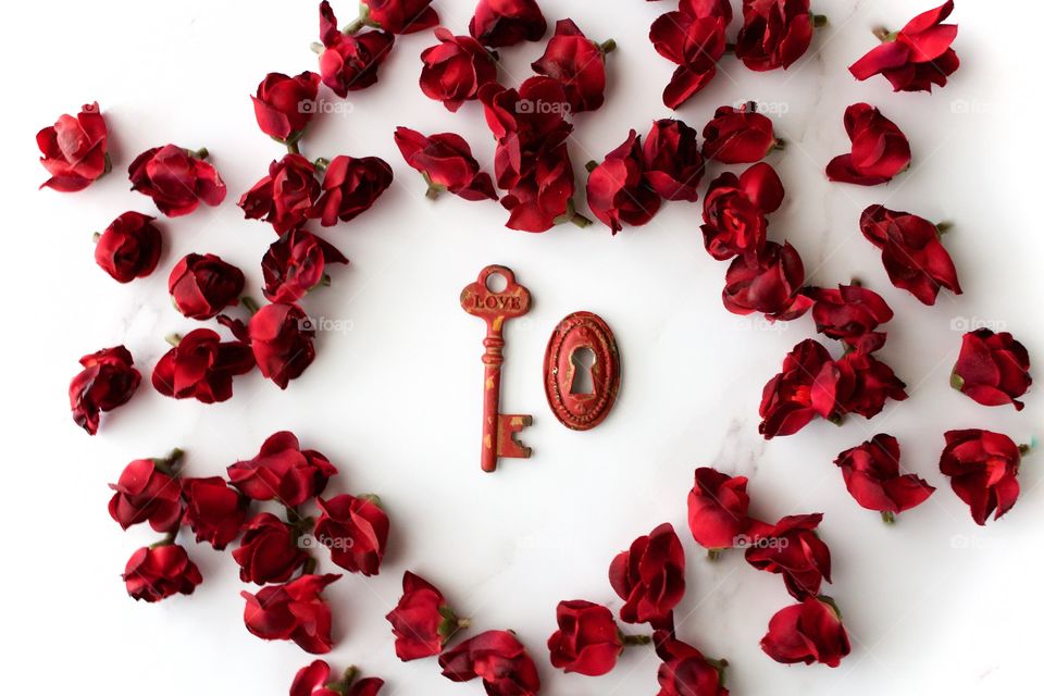 Red, metal, vintage key and keyhole with red, silk rose buds arranged in hear shape on white marble surface
