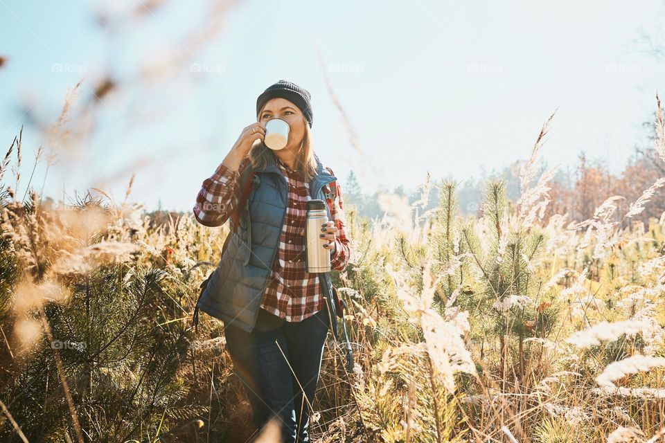 Woman enjoying the coffee in bright warm sunlight during vacation trip. Woman standing in tall grass and looking away holding cup of coffee and thermos flask. Woman with backpack hiking along path in mountains. Active leisure time close to nature