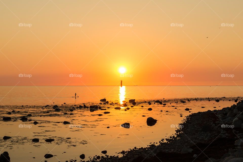 One lonely stand up paddler and the silhouette of a lighthouse at peaceful orange colored sunset 