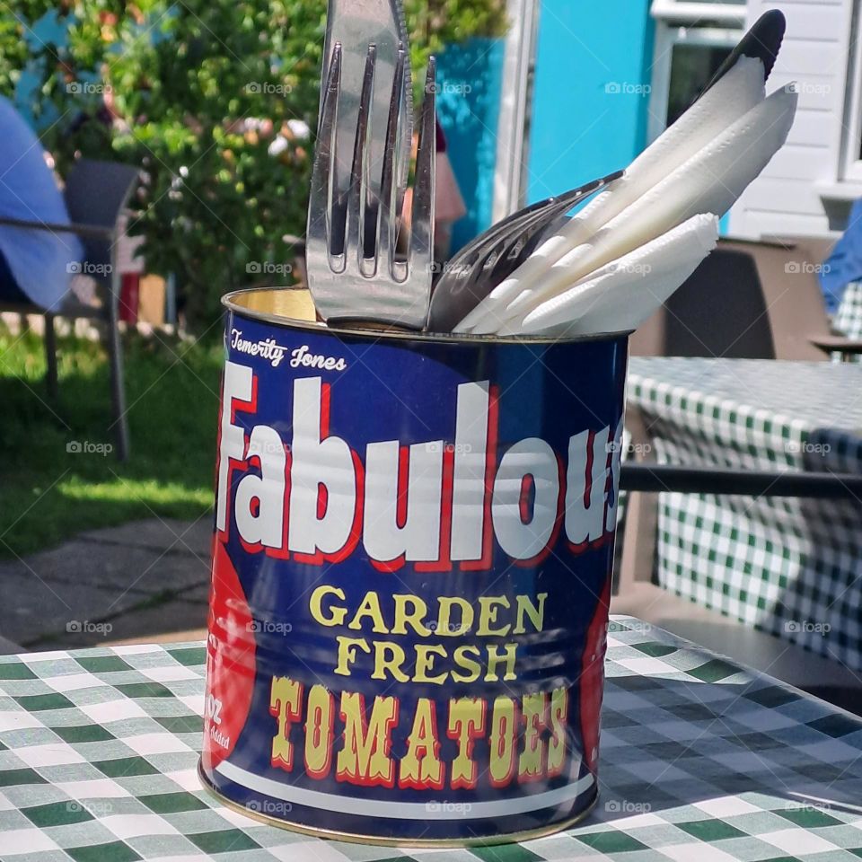 restaurant garden. cutlery and napkins in a large used tin. checked tablecloth outside sunny