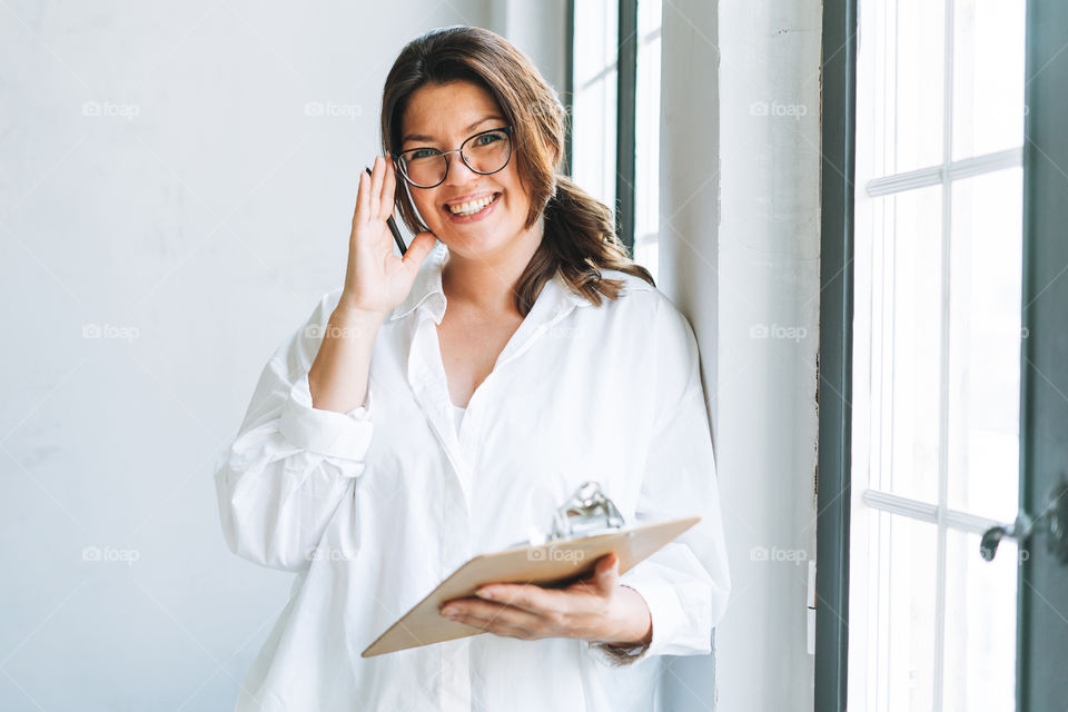 Young smiling brunette woman plus size in white shirt near window with pen and documents in hands in bright modern office