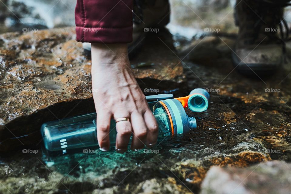 Woman taking pure water to bottle from mountain stream during trekking in mountains. Hiker crouching on rocks, filling bottle up with cold mountain water. Enjoying the outdoors in the summer trip vacation