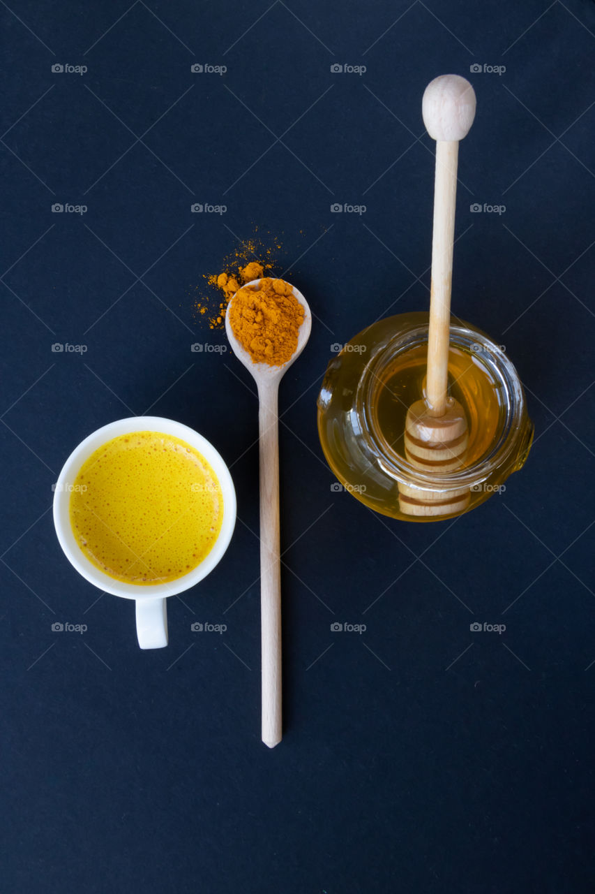 Top view to a cup with golden milk, some turmeric in the wooden spoon and honey in the jar.