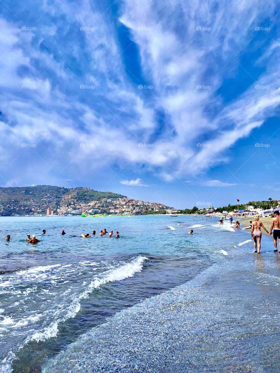 Photo with the beach, sea and mountains in the background. Many happy people swim in the sea, creating an atmosphere of fun and carefree relaxation.
