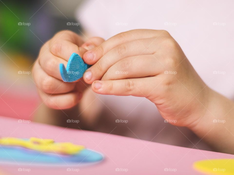 Hands of little caucasian girl peel off paper with felt sticker blue hat for crafts,sitting at the table,side view close-up. Creative kids concept, Easter preparation.