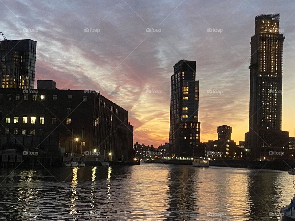 Newtown Creek in LIC, Queens, New York with the sun setting behind the horizon giving the cloudy overcast sky a golden glow. The dark silhouettes of several high rise buildings frame the composition in this picture. 2021. Hypnotic Productions