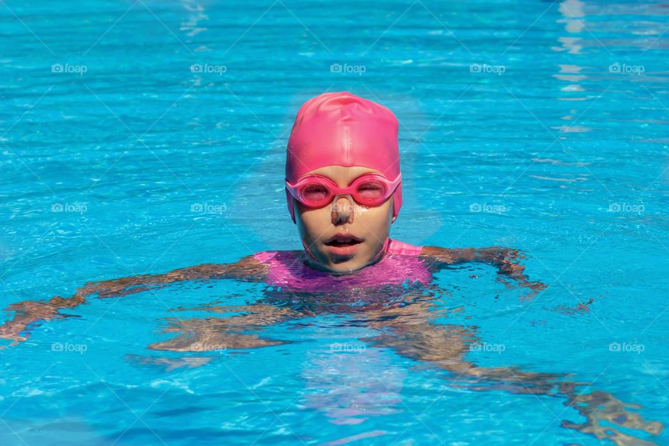 Child in swimming cap, goggles and nose clip in swimming pool looking at camera