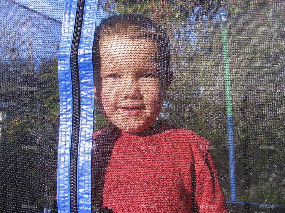 A Little boy takes a moment and stops jumping on the trampoline. He's happy to be photographed as he stands still behind the textured safety nets.