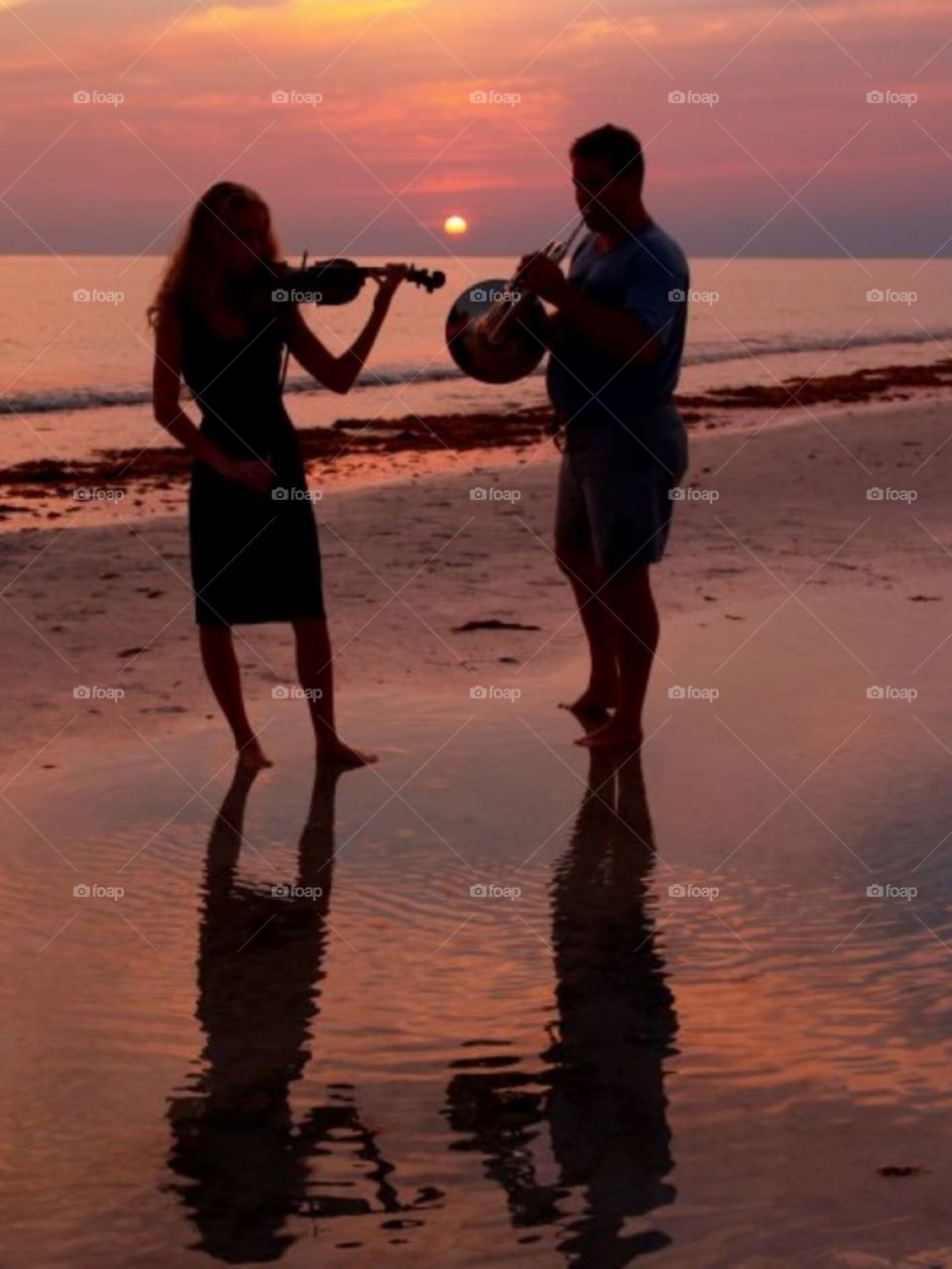 Violinist and French horn musician playing sweet music by the ocean on the beach as the sun sets behind them 