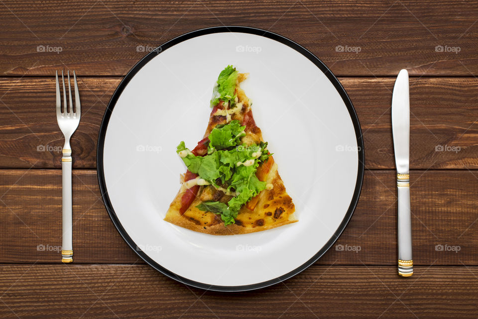 A piece of pizza on white plate with a knife and fork on wooden background